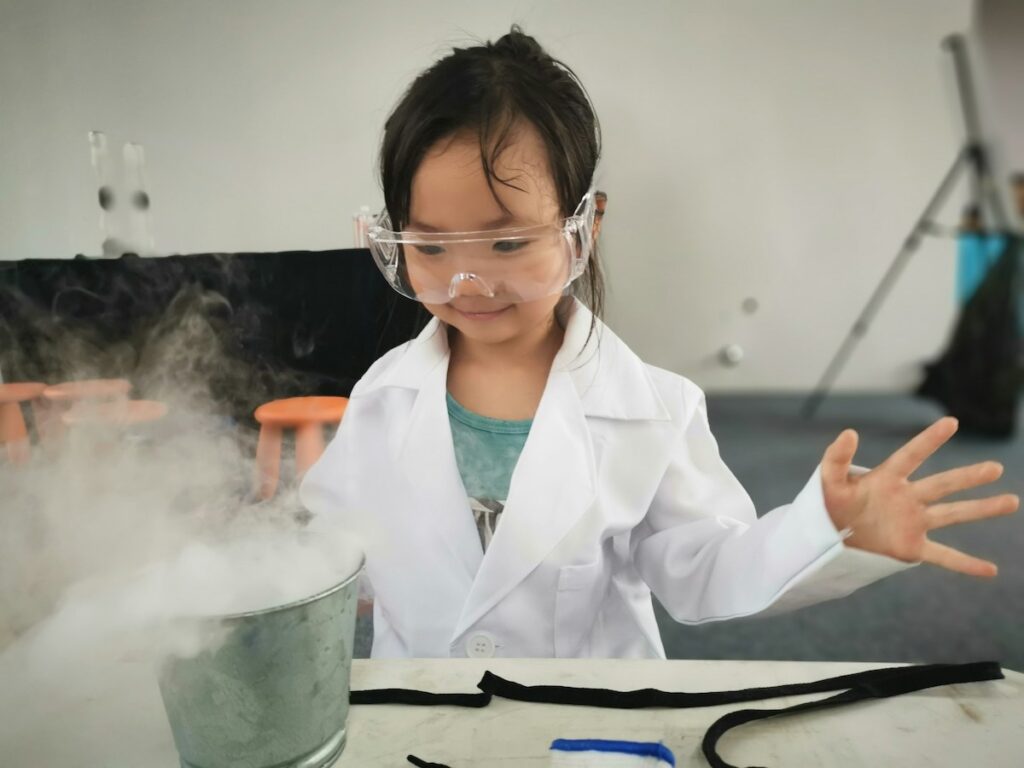 Ein Kind in weißem Laborkittel und Schutzbrille experimentiert fasziniert mit rauchenden Flüssigkeiten, passend zum Gewinnspiel für die Wissenschaftsstunde in Wien, kuratiert auf fini.wien.