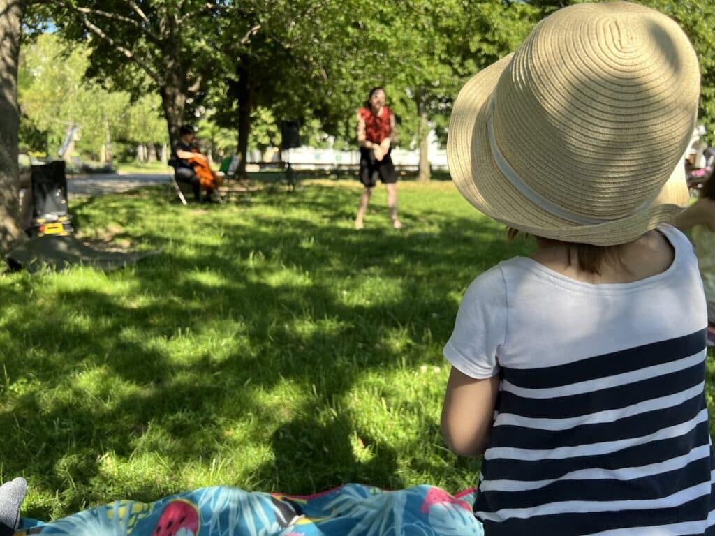 Ein Kind mit hellem Strohhut sitzt auf einer Picknickdecke im grünen Park und schaut gespannt einer Theateraufführung unter freiem Himmel zu. Ein Beispiel für kostenlose Openair-Highlights in Wien, kuratiert für Familien auf fini.wien.